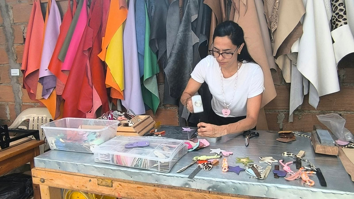 A woman leather master is crafting a bag with hand tools at her small leather atelier with hanging colorful pieces of leather in the background